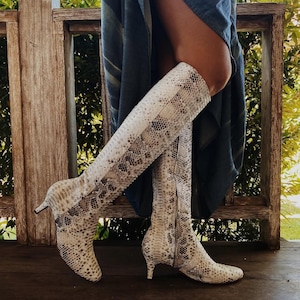 May include: A pair of knee-high boots featuring a snakeskin pattern in shades of cream, beige, and dark brown. The boots have a pointed toe, a slim kitten heel, and a full-length side zip closure, extending up to the lower portion of the knee.