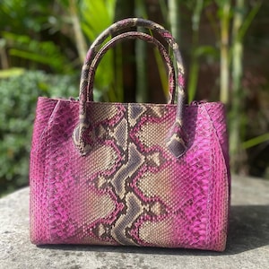May include: A structured handbag featuring a vibrant magenta and natural tan snakeskin pattern. The bag has two matching top handles and a rectangular body with a distinct scale texture that transitions from dark pink sides to a central tan panel.