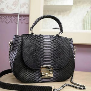 May include: A dark grey and black handbag featuring a textured snakeskin pattern. The bag has a structured top handle, a silver-toned metal clasp closure on the front flap, and a detachable shoulder strap made of a matching chain and leather material.
