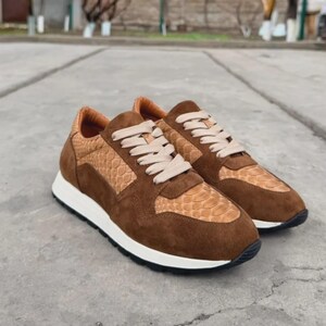 May include: A pair of low-top sneakers featuring a combination of brown suede and tan snakeskin-textured panels. The shoes include beige laces, a white midsole, and a black outsole, showcasing a layered design with a rounded toe and padded collar.