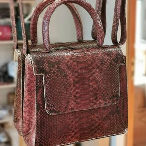 Könnte beinhalten: Eine strukturierte Handtasche mit einem dunkelroten und schwarzen Schlangenledermuster. Die Tasche hat einen rechteckigen Korpus, einen Überschlag, zwei abgerundete Tragegriffe und zwei lange vertikale Riemen an der Oberseite zum Tragen.