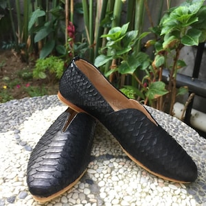 Könnte beinhalten: Ein Paar schwarze Slipper mit einem strukturierten, schuppenartigen Muster auf der Außenseite. Die Schuhe haben eine spitz zulaufende Form, einen tief ausgeschnittenen Vorderbereich mit V-Ausschnitt und eine hellbraune Sohle.