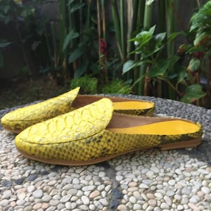 May include: A pair of bright yellow slip-on mules featuring a textured snakeskin pattern on the exterior. The shoes have a pointed toe, a flat sole, and a smooth yellow interior lining. The design is backless, allowing for easy entry and exit.