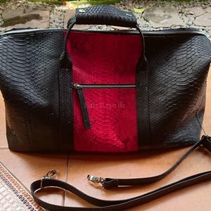 Op de afbeelding: Een reistas met een zwart en rood slangenleer-textuurpatroon. De tas heeft een centraal rood paneel met een ritsvak, zwarte zijpanelen, een handvat aan de bovenkant en een afneembare zwarte schouderriem. De tekst Faiz Reptile is zichtbaar.