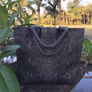 May include: A dark grey tote bag featuring a textured snakeskin pattern across the entire surface. The bag has two matching handles and a central vertical panel detail. The material displays a consistent scale-like appearance throughout.
