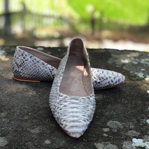 May include: A pair of pointed-toe flats featuring a textured grey and white snakeskin pattern. The shoes have a low-profile silhouette with a tan-colored sole and a smooth, light-colored interior lining visible within the opening of the footwear.