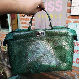 May include: A structured handbag featuring a green and black snakeskin-textured exterior. The bag includes a single top handle, a silver-toned metal turn-lock closure on the front flap, and side gussets that extend slightly beyond the main body.