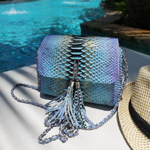 May include: A rectangular shoulder bag featuring a textured, iridescent snakeskin-patterned finish in shades of blue, purple, and gold. The bag includes a silver-toned chain strap and a prominent central tassel ornament with matching metallic accents.