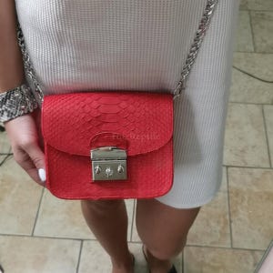 May include: A bright red crossbody bag with a textured snakeskin pattern and a silver-toned metal clasp closure. The bag features a silver chain shoulder strap and a rectangular flap design. The text Fat Reptile is embossed on the front flap.