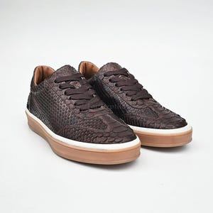 May include: A pair of low-top sneakers featuring a dark brown snakeskin-textured upper, matching dark brown laces, a white midsole, and a translucent gum-colored rubber outsole. The design includes a rounded toe and a padded collar.