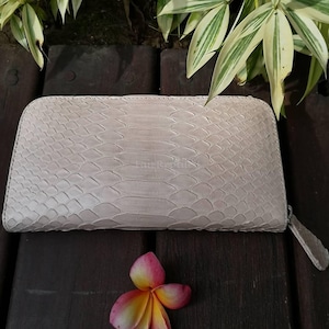 Peut inclure: Un portefeuille rectangulaire présentant une texture en peau de serpent beige clair avec un motif d'écailles répétitif. Le portefeuille a un design aux bords arrondis et comprend une tirette de fermeture éclair beige assortie.
