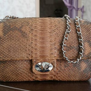 Pode incluir: Uma bolsa de ombro retangular com um padrão de pele de cobra em tons de castanho e bronzeado. A bolsa possui uma alça de corrente de metal prateado e um fecho giratório prateado na aba frontal. A textura das escamas é visível na superfície.