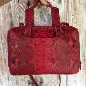 Könnte beinhalten: Eine rechteckige Handtasche mit einem leuchtend roten und schwarzen Schlangenledermuster. Die Tasche verfügt über zwei Tragegriffe, einen Reißverschluss an der Oberseite und einen abnehmbaren Schulterriemen. Die Oberfläche zeigt das Schuppenmuster.