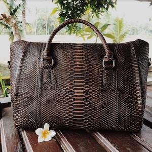 May include: A dark brown handbag featuring a textured snakeskin pattern across the entire exterior. The bag has two sturdy, braided handles attached with silver-toned metal hardware. A single white and yellow flower rests in the foreground.