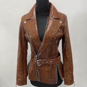 Könnte beinhalten: Eine braune Jacke mit schlangenlederartiger Textur, ausgestattet mit einem Reverskragen, langen Ärmeln und einem silberfarbenen diagonalen Frontreißverschluss. Das Kleidungsstück verfügt über einen passenden Gürtel mit silberner Schnalle.