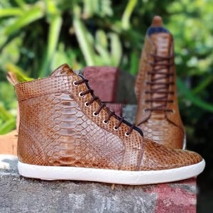 May include: A pair of high-top sneakers featuring a brown snakeskin-textured pattern. The shoes have dark brown laces threaded through silver-toned metal eyelets and are finished with a solid white rubber sole that wraps around the base.