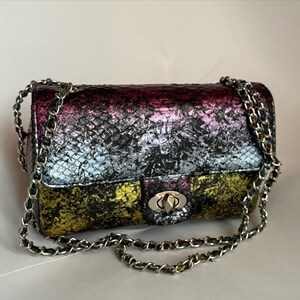 Puede incluir: Un bolso de hombro rectangular con un patrón de piel de serpiente texturizado en un degradado de tonos metálicos morados, azules y amarillos. El bolso incluye una correa de cadena de metal plateado y un cierre giratorio plateado en la solapa.