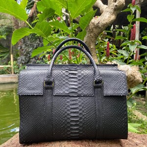 May include: A structured black handbag featuring a distinct snakeskin texture across the entire exterior. The bag includes two top handles, a rectangular main body, and a flap closure that extends over the front, showcasing a consistent scale pattern.