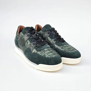 Pode incluir: Um par de ténis verde-escuro com um topo texturizado com padrão de pele de cobra combinado com painéis de camurça verde-escuro liso. Os sapatos têm atacadores pretos, forro interior bege e uma sola de borracha branca espessa e plana.