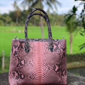 May include: A structured tote bag featuring a pink and black snakeskin pattern. The handbag has two matching top handles with a textured scale design. The body of the bag displays a vertical central panel with a distinct scale alignment, flanked by side panels with a darker, irregular snakeskin print.