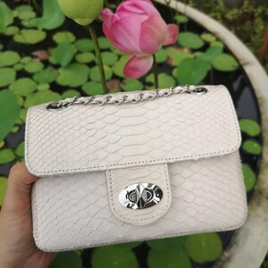 May include: A cream-colored handbag with a textured snakeskin pattern, featuring a silver-tone metal chain strap and a polished silver-tone oval twist-lock closure on the front flap. The bag has a structured rectangular shape and stitching.