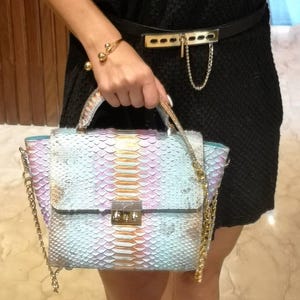 Puede incluir: Un bolso estructurado con un exterior de patrón de piel de serpiente multicolor en tonos azul pastel, rosa y dorado. El bolso incluye un asa superior, una correa de hombro de cadena dorada desmontable y un cierre metálico rectangular.
