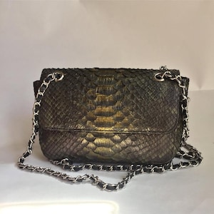 Puede incluir: Un bolso de hombro rectangular con un patrón de textura de piel de serpiente en tonos bronce metálico oscuro y negro. El bolso incluye un cierre de solapa y una correa de cadena de metal plateado entrelazada con material negro.