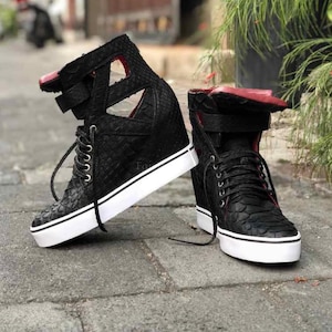 Puede incluir: Un par de zapatillas de cuña altas de color negro con un patrón texturizado similar a la piel de serpiente, suelas de goma blanca, cordones negros y forro interior rojo. El calzado presenta un diseño con aberturas en el tobillo.