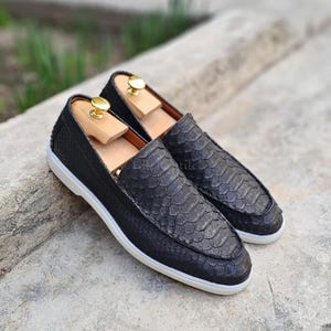 May include: A pair of black loafers featuring a textured snakeskin-patterned exterior, a low-profile silhouette, and a white rubber sole. Each shoe includes a light-colored wooden shoe tree with a gold-toned metal knob inserted inside the opening.
