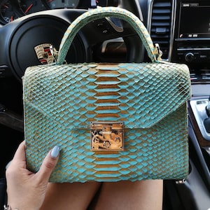 May include: A structured handbag featuring a textured, scale-like pattern in alternating shades of turquoise and metallic gold. The bag includes a top handle, a fold-over flap, and a polished gold-tone rectangular metal clasp closure at the center.