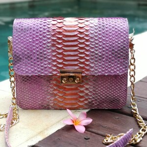 May include: A rectangular shoulder bag featuring a textured snakeskin pattern with a gradient colour transition from purple on the sides to a vibrant orange centre. The bag includes a gold-tone metal clasp closure and a matching gold chain strap.