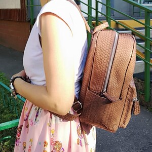May include: A brown backpack with a textured snakeskin-like pattern, featuring a silver zipper closure, a top handle, and adjustable shoulder straps. The bag has a structured rectangular shape with a smaller front pocket and matching brown zipper pulls.