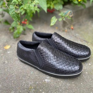 May include: A pair of black slip-on shoes featuring a textured, scale-like pattern across the upper surface. The shoes have elastic side panels for a snug fit and a solid black sole with a thin white stripe running along the edge of the base.
