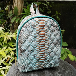 May include: A small backpack featuring a textured snakeskin pattern in shades of light blue, brown, and grey. The bag has a rounded top, a top handle, and a bright teal zipper closure that runs along the perimeter of the main compartment.