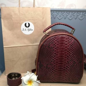 May include: A deep burgundy, textured backpack with a top handle and gold-tone zip. Beside it sits a matching burgundy cuff bracelet, a white plumeria flower, and a small surfboard-shaped keyring with a sunset graphic, all near a paper bag.