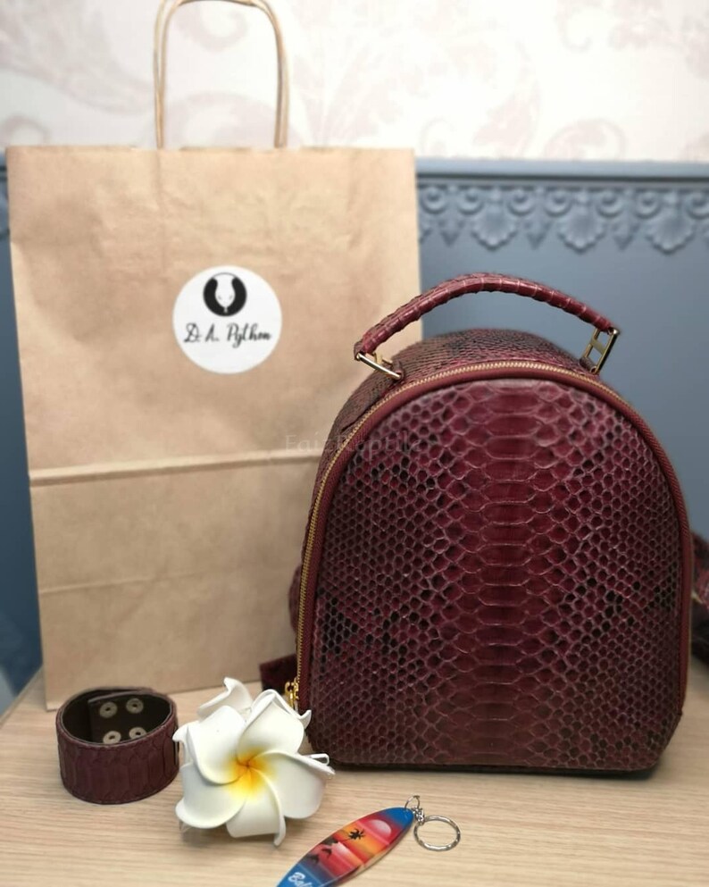 K&ouml;nnte beinhalten: Ein tiefburgunderfarbener, texturierter Rucksack mit Tragegriff und goldfarbenem Rei&szlig;verschluss. Daneben liegen ein passendes burgunderfarbenes Armband, eine wei&szlig;e Plumeria-Bl&uuml;te und ein kleiner surfbrettf&ouml;rmiger Schl&uuml;sselanh&auml;nger.