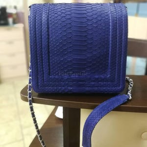 Puede incluir: Un bolso de hombro rectangular de color azul vibrante con un patrón de piel de serpiente texturizado y un cierre de solapa estructurado. El bolso incluye una correa de cadena de metal plateado con una hombrera de piel de serpiente azul y el texto FaizReptile.
