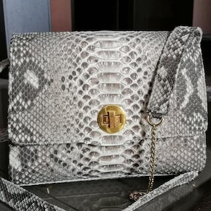 May include: A rectangular shoulder bag featuring a grey and white snakeskin pattern. The bag has a gold-toned circular turn-lock closure at the centre of the front flap. It includes a matching snakeskin strap and a thin gold-toned metal chain detail.