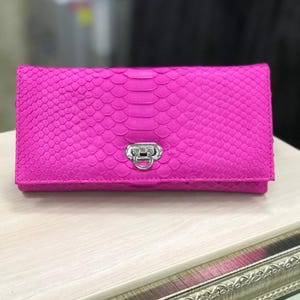 May include: A vibrant hot pink rectangular clutch wallet featuring a textured snakeskin pattern. The wallet has a fold-over flap secured by a polished silver-toned metal turn-lock closure centered on the front, providing a secure fastening mechanism.