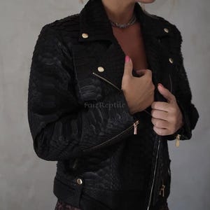 Könnte beinhalten: Eine schwarze Motorradjacke mit einem strukturierten Schlangenledermuster. Die Jacke verfügt über goldfarbene Metallbeschläge, darunter Reißverschlüsse an der Vorderseite und den Ärmeln, Druckknöpfe an den Revers und einen Gürtel mit Schnalle in der Taille. Der Text FaizReptile ist sichtbar.