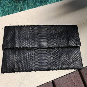 May include: A rectangular black clutch bag featuring a textured snakeskin pattern. The design includes a central vertical panel with larger, elongated scales flanked by two side sections with smaller, uniform scales, creating a distinct visual contrast.