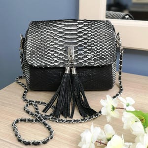 Puede incluir: Un bolso bandolera rectangular con una solapa de textura de piel de serpiente en plata y negro sobre una base negra sólida. El bolso incluye una correa de cadena metálica plateada entrelazada con material negro, un cierre central y dos borlas.