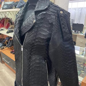 May include: A black jacket featuring a textured, scale-like pattern across the entire surface. The garment includes a notched collar, a full-length silver-tone front zip, two zipped side pockets, and a buckled belt at the hem. The text FaizReptile is visible.