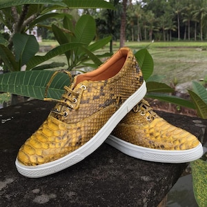 May include: A pair of low-top sneakers featuring a yellow and brown snakeskin-patterned exterior, white rubber soles, and matching yellow laces threaded through silver-toned metal eyelets, with a tan interior lining visible at the collar.