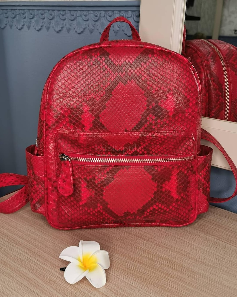 May include: A bright red backpack featuring a dark red snakeskin pattern throughout. The bag includes a top handle, adjustable shoulder straps, two side pockets, and a front zippered compartment with the text PaizReptile printed on it.