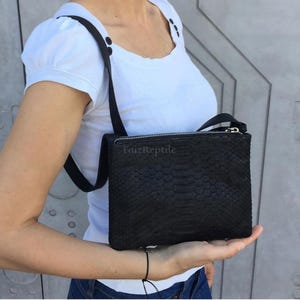 May include: A black rectangular crossbody bag with a textured snakeskin pattern and a silver zip closure. The bag features a thin black shoulder strap and has the text FaizReptile printed in small, subtle letters on the front surface.