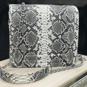 Peut inclure: Un sac à bandoulière rectangulaire présentant un motif peau de serpent gris, blanc et noir. Le sac possède une fermeture à rabat structurée et une bandoulière en chaîne métallique argentée avec une épaulette assortie au motif serpent.