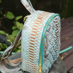 May include: A small backpack featuring a textured snakeskin pattern in shades of light blue, silver, and tan. The bag includes a bright teal zipper closure with gold-toned hardware, a top handle, and adjustable shoulder straps in matching material.