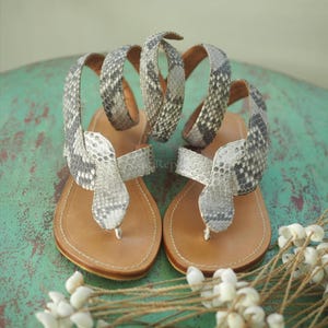 May include: A pair of flat thong sandals featuring a snake-shaped strap design with a grey and white snakeskin pattern. The sandals have a tan leather footbed with visible stitching along the edges and long, wrap-around ankle straps.