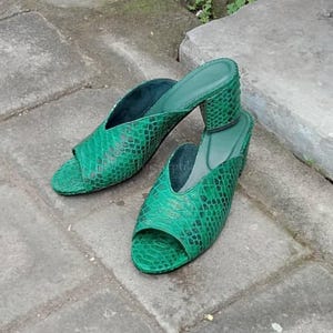 May include: A pair of emerald green open-toe mules featuring a textured snakeskin pattern. These slip-on shoes have a low block heel and a V-shaped vamp, showcasing a consistent green scale design across the entire exterior surface of the footwear.